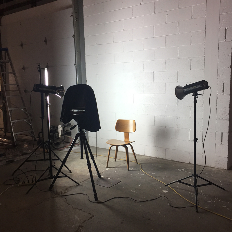 A white cement wall with a single empty chair illuminated with studio lights and large format camera